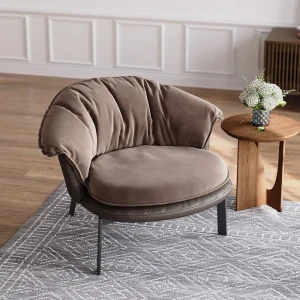 Taupe suede accent chair with rounded back and black legs, styled beside a sculptural wooden side table with white flowers in a glass vase, set on a gray geometric rug.