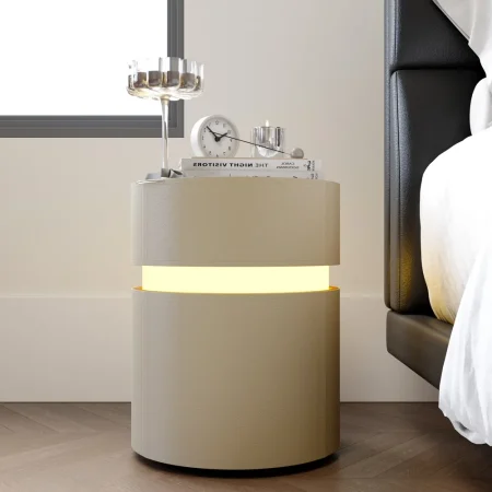 Cylindrical beige nightstand with glowing light band, styled with books, clock, and decor beside a modern bed.