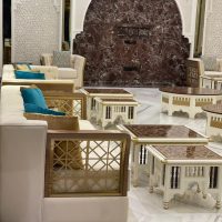 A regal lounge composition blending Islamic architectural motifs, sculptural furnishings, and rich material contrasts. White and gold sofas with turquoise cushions, marble-top tables, red marble wall with arched niche and carvings.
