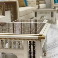 A refined living room vignette blending traditional craftsmanship, plush seating, and luminous material contrast. White carved coffee table with glossy patterned top, beige sofas with blue and gold cushions, marble flooring.