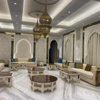A culturally immersive lounge ensemble blending Islamic architectural elegance, sculptural furnishings, and ambient lighting. White and gold sofas with turquoise cushions, geometric coffee tables, arched wall carvings, and hanging lanterns.
