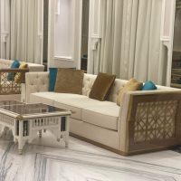 A formal lounge ensemble blending plush symmetry, carved elegance, and reflective architectural detailing. Cream tufted sofas with gold and blue cushions, carved white coffee table, marble floor, and mirrored wall panels.