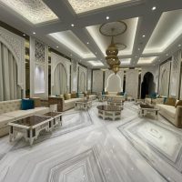 A grand lounge ensemble blending Islamic architectural symmetry, plush seating, and layered marble geometry. Beige sofas with turquoise cushions, white carved tables, arched wall panels, gold chandeliers, and marble flooring.
