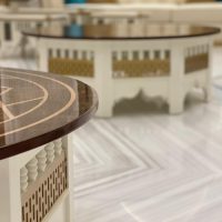 A sculptural table duo blending geometric inlay, spindle craftsmanship, and luminous material contrast. Two round tables with dark wood tops, white carved bases, gold lattice accents, and marble flooring.