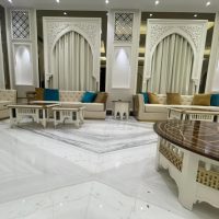 A symmetrical lounge ensemble blending Islamic architectural elegance, plush seating, and geometric marble textures. Beige tufted sofas with teal cushions, white carved coffee tables, arched wall niches, and marble flooring.