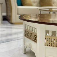 A richly detailed lounge vignette blending artisanal craftsmanship, plush textures, and luminous material contrast. White carved coffee table with dark wood inlay, beige tufted sofa with gold and teal cushions, marble flooring.