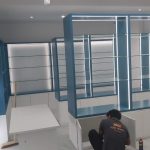 Clean lines and illuminated shelving define this retail space in progress, blending function with sleek design. Modern retail interior with blue-framed glass shelving units, white base cabinets, LED lighting, and white tiled flooring in a minimalist setup.