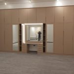 A refined wardrobe design blends symmetry and function with a central vanity and ambient lighting. Built-in wardrobe with symmetrical cabinets, central vanity area with mirror and lighting, grey flooring, and white ceiling in a modern interior.