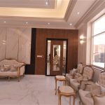 Classical charm meets modern luxury in this sunlit living space with gold accents and refined textures. Elegant living room with white and gold tufted furniture, marble flooring, decorative wall panels, and large windows in a classical European-style interior.