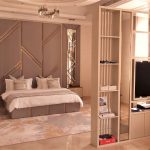 A multifunctional layout blends elegance and entertainment in this refined bedroom retreat. Luxurious bedroom with padded geometric headboard, white linens, partition wall with built-in shelving and TV, and chandelier lighting in a modern interior.