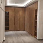 Warm wood tones and ambient lighting bring depth and elegance to this contemporary dressing space. Walk-in closet with natural wood cabinetry, open shelving, recessed tray ceiling lighting, and dark gray flooring in a minimalist modern interior.