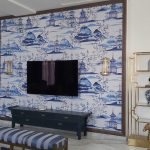 A fusion of traditional motifs and modern luxury brings depth and harmony to this blue-and-gold living space. Elegant living room with blue-and-white mural, wall-mounted TV, gold shelving units, dark blue console, and striped bench in a modern Asian-inspired interior.