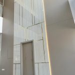 “Hidden door + marble + brass grid = the ultimate ‘wow’ before you even say hello.” A dramatic double-height entrance feature wall clad in large light beige marble slabs with subtle veining, accented by polished brass inlays forming an asymmetric geometric grid. A concealed matte taupe door with a single vertical brass strip is seamlessly integrated, illuminated by warm hidden LED lighting from the side.