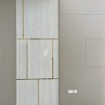 “Marble + polished brass inlays = quiet luxury that speaks volumes.” A tall modern feature wall panel clad in large rectangular slabs of light beige veined marble, divided by thin horizontal and vertical polished brass inlays forming an asymmetric geometric pattern. The panel is flanked by matte taupe/gray wall sections with concealed doors or access panels, set above glossy white marble flooring.