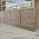 “Travertine patchwork + brass inlays = quiet luxury just got loud.” A luxurious modern sideboard with four doors covered in an irregular geometric patchwork of light travertine-look panels outlined in thin brushed-brass inlays. A large gold-framed geometric mirror hangs above, set against a cream wall with built-in shelving, on glossy white marble flooring.