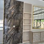 “Book-matched marble + gold geometric mirror + 3D tiles = quiet luxury overload.” A luxurious modern feature wall combining dramatic book-matched dark marble with swirling brown, gray, and gold veining on the left, a recessed cream-painted shelving niche in the center, and a large geometric gold-framed mirror reflecting the room on the right. Below the mirror, a textured 3D-patterned cabinet in cream and gold finishes with brass trim.