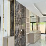 “Book-matched marble + brass everything = quiet luxury just got loud.” A luxurious modern entryway or hallway featuring a dramatic full-height book-matched dark marble wall with swirling brown, black, and gold veining edged in brass. A recessed cream shelving niche sits beside a large geometric gold-framed mirror above a textured 3D tiled sideboard in cream and brass tones, all on polished white marble flooring.