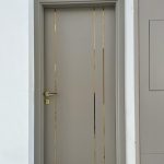 “Taupe + triple brass stripes = the chicest door you’ll ever walk through.” A sleek modern interior door in matte taupe-gray with three full-height vertical brushed-gold metal inlays, framed in crisp white with a minimalist gold lever handle.