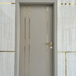 “Taupe + brushed brass inlays = the most stylish door you’ll ever close.” A sleek modern interior door in matte taupe-gray with three vertical inlaid brushed-gold metal strips running full height, set into a light marble wall accented by thin horizontal and vertical gold inlays.
