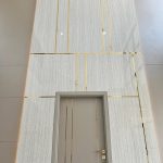 “Hidden door + marble + brass inlays = quiet luxury on steroids.” A dramatic double-height wall clad in large panels of light beige marble with subtle veining, accented by thin polished brass inlays forming an asymmetric geometric grid. A recessed matte taupe door with three vertical brass strips is perfectly integrated into the design, creating a seamless luxurious feature wall.