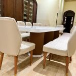 “Cream quilted leather + glossy white + walnut & gold = dinner just got royal.” A luxurious formal dining room featuring a long oval high-gloss white dining table with dramatic quilted walnut pedestal bases, surrounded by ten cream quilted-leather dining chairs and one matching bench, all with walnut legs and gold foot caps. A large dark-wood china cabinet displays white porcelain in the background.