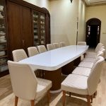 “Cream quilted chairs + glossy white table + walnut & gold details = dinner just got royal.” A luxurious formal dining room with a long oval white-gloss dining table seating twelve, surrounded by cream quilted-leather high-back chairs with walnut legs and gold foot caps. A built-in dark-wood display cabinet filled with white china lines one wall, illuminated by a traditional ornate chandelier.