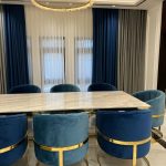 A refined dining room ensemble blending jewel-toned seating, sculptural lighting, and polished marble elegance. White marble dining table with blue and teal chairs, gold frames, crystal chandelier, and layered curtains.