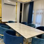 A sophisticated dining ensemble blending plush seating, sculptural lighting, and elegant architectural framing. Marble dining table with gold legs, blue velvet chairs, crystal chandelier, and white-paneled walls with curtains.