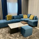 A vibrant lounge composition blending bold upholstery, soft textures, and minimalist architectural framing. Blue sectional sofa with yellow cushions, marble coffee table, blue ottoman, grey rug, and layered curtains.