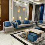 A refined living room ensemble blending plush textures, layered lighting, and bold accent contrasts. Light gray tufted sofas with blue pillows, marble coffee tables, blue ottomans, geometric rug, and crystal chandelier.