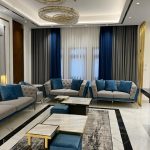 A grand living room ensemble blending plush seating, layered textiles, and opulent lighting. Tufted grey and blue sofas with marble coffee tables, abstract rug, layered curtains, and crystal chandeliers.