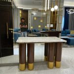 A luxurious entryway composition blending reflective surfaces, sculptural lighting, and rich material contrast. Console table with marble top, mirrored wall, gold partition, marble flooring, and blue-gray seating area.