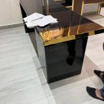 A sleek reception desk composition blending reflective surfaces, active workspace cues, and minimalist elegance. Glossy black desk with gold trim, open notebook with highlighted text, and light wood flooring.