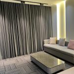 A minimalist lounge composition blending soft neutrals, ambient lighting, and geometric surface layering. Light gray sofa with pastel cushions, marble coffee table, geometric carpet tiles, and vertical curtain backdrop.