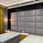 A symmetrical twin bedroom setup blending padded textures, warm lighting, and vibrant accent tones. Twin beds with grey padded headboard, yellow-accented nightstand, wall lights, and grey-yellow curtains.