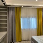 Layered textures and soft lighting bring depth and elegance to this contemporary bedroom design. Modern bedroom with layered curtains, built-in shelving, recessed ceiling lighting, and a bed with protective plastic in a sleek interior.