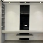 Sleek symmetry and neutral tones define this contemporary setup designed for focus and function. Minimalist interior with wall-mounted desk, glossy black panel, built-in shelving units, and recessed ceiling lighting in a modern workspace.