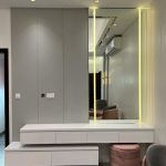 Sleek lighting and soft textures elevate this console setup into a refined design statement. Modern vanity area with illuminated mirror, white staggered drawers, pink quilted ottoman, and grey walls in a stylish interior.