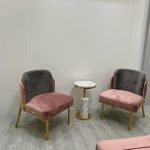 Chic contrasts and elegant finishes define this stylish corner setup. Modern seating area with gold-framed chairs, pink and gray upholstery, marble side table, and minimalist white walls in a contemporary interior.