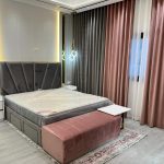 Soft hues and layered textures bring elegance and warmth to this contemporary bedroom. Stylish bedroom with dark grey upholstered headboard, pink bench, white nightstands, and dual-tone curtains in a modern interior.