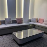 A contemporary lounge layout blending soft geometry, muted tones, and vertical ambient illumination. L-shaped grey sectional sofas with throw pillows, marble coffee table, striped carpet tiles, and ambient wall lighting.