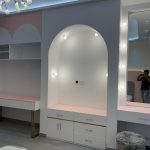 A playful yet elegant vanity zone blending soft hues, built-in lighting, and functional display geometry. Vanity and storage area with white and pink cabinetry, gold accents, lighted mirror, and decorative arches.