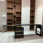 A refined workspace layout blending sculptural furniture, warm lighting, and minimalist architectural styling. Black desk with gold accents, wooden bookshelves, floor lamp, side table, and light wood flooring.