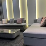 Soft textures and warm lighting create a cozy yet refined lounge atmosphere. Contemporary living room with L-shaped gray sectional sofas, marble coffee table, vertical panel lighting, and dark curtains in a modern interior.