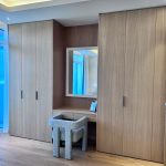 “Oak everything + bouclé chair + hidden lighting = quiet luxury getting-ready goals.” A sleek built-in bedroom vanity nook flanked by full-height light-oak wardrobes. A floating wood vanity with a rectangular illuminated mirror sits centrally, paired with a plush gray-bouclé curved chair. Recessed ceiling lighting and hidden LED strip lighting create a soft, luxurious glow.