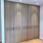 “Light oak + fluted details + zero visible handles = closet perfection.” A sleek, full-wall built-in wardrobe with six tall doors in light natural oak veneer, featuring minimalist recessed handles and subtle vertical fluted detailing on the lower panels of the center doors. Warm recessed ceiling lights highlight the wood grain, set against light wooden flooring.