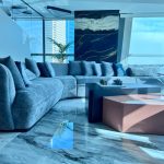 “Curved gray cloud sofa + black & pink table + that insane abstract art = penthouse perfection.” A luxurious high-rise living room with panoramic city skyline views through floor-to-ceiling windows. A large curved light-gray plush sectional sofa faces a modern two-tier coffee table in matte black and glossy rose-pink, set on high-gloss white marble flooring. A dramatic oversized abstract artwork in black, gold, and white hangs above.