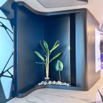 “Matte-black pod + glowing tropical glow-up = the most extra planter in existence.” A futuristic curved matte-black architectural alcove featuring a built-in illuminated planter with a large artificial banana-leaf plant and monstera surrounded by white pebbles, dramatically lit from above against glossy white marble flooring in a high-tech luxury interior.