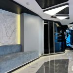 “Powder-blue velvet + glowing marble waves = quiet luxury just went full sci-fi.” A futuristic luxury lounge corner featuring a long built-in light-blue velvet bench set against a dramatically backlit wavy marble wall panel. Hidden warm LED strips glow beneath the bench and along the base, while angular black geometric shelving with integrated lighting sits to the right, all on high-gloss marble flooring under a sculptural ceiling with triangular light fixtures.