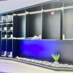 “Matte black + blue glow + hidden planter = showroom flex on another level.” A sleek futuristic black reception or display counter with integrated open shelving on the left, illuminated by warm LED strips. A built-in planter with white pebbles and a small green succulent sits at the base, accented by dramatic blue under-counter lighting.